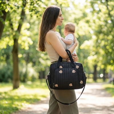 sac a langer bebe accroché au bras d'une maman portant bébé dans les bras au parc